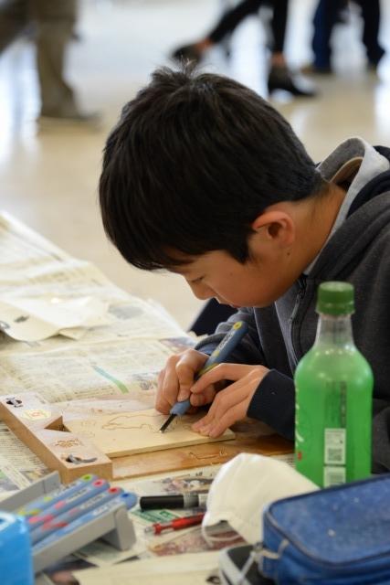 水色の彫刻刀を使って、木の板を削っている少年の写真