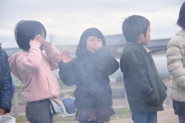 焚き火の煙の中、目や鼻に手を当てて避けている子ども達の写真