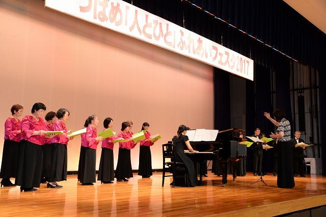壇上でピアノ演奏している女性と指揮者に合わせて合唱してるピンクの服と黒のスカート姿の女性たちの写真