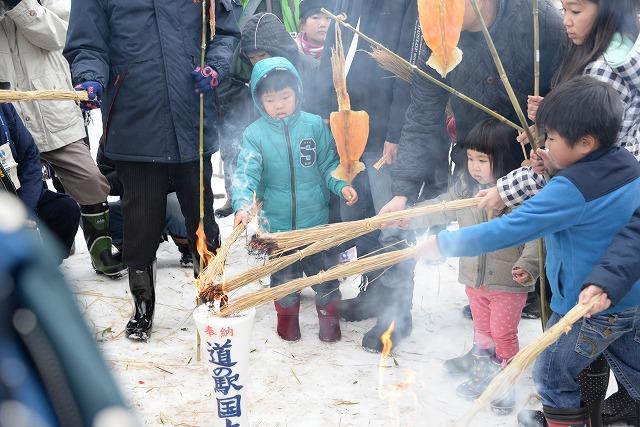 大きいろうそくに近づいて長い藁に火を近づけ点火する3人の幼児たちの写真