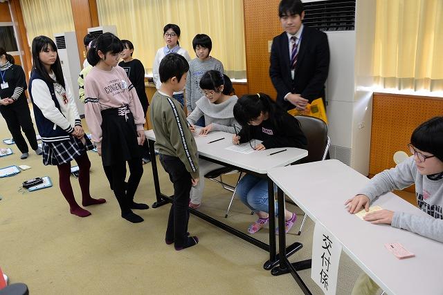 2つの白いテーブルを並べて選挙受付の体験をしている女子生徒とその後にいる男性職員と列を作って待つ生徒たちの写真