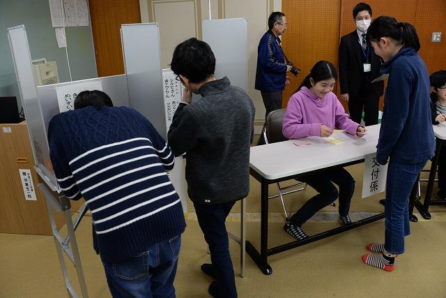 投票用紙を交付した生徒が本物の記載台で用紙に候補名を書いている様子と交付係の女子生徒の写真