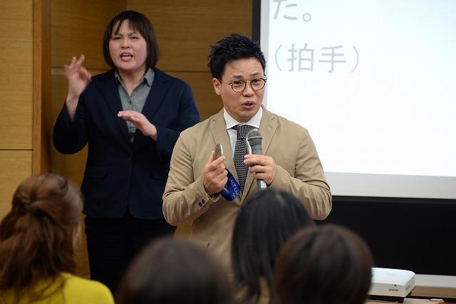 清水宏保氏が持参した銀メダルを披露している様子とその後で手話通訳をしている女性の写真
