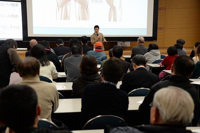 講演をする清水宏保氏と多くの参加者たちを会場の後ろから撮影した写真
