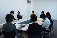 白い壁に囲まれた部屋で四角に並べられた机を囲みながら資料を見つつ会議に参加する女性たちの写真