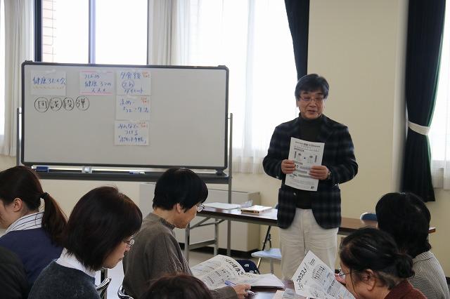 ホワイトボードの隣で資料を両手に持ち講義する志賀誠治先生とその聞いている参加者立ちの写真