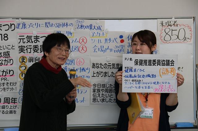 プロジェクト「保健推進協議会」の活動内容をマイクを持って発表する女性参加者と紙を掲げる女性職員の写真