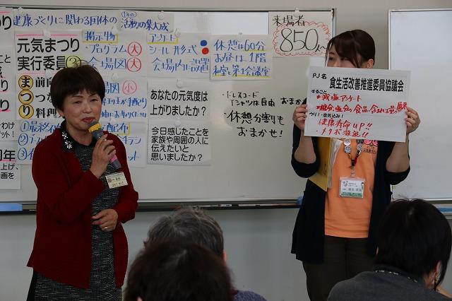 プロジェクト「食生活改善推進委員協議会」の活動内容をマイクを持って発表する女性参加者と紙を掲げる女性職員の写真