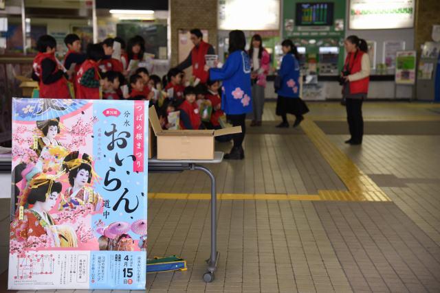 分水花魁道中のポスターとはっぴを着た女性と観光パンフレットを手に集まっている小学生たちの写真