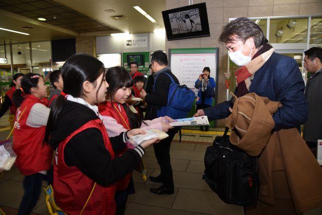 東京からの新幹線の乗客にお米と観光パンフレットを手渡す小学生たちの写真