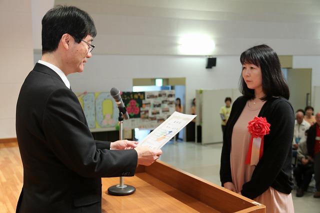 「つばめ元気かがやきポイント事業表彰・認定式」で、受賞者の女性に鈴木市長がマイクに向かって表彰状を読んでいる写真