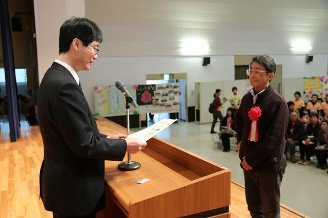 受賞者の男性に鈴木市長がマイクに向かい表彰状を読んでいる写真