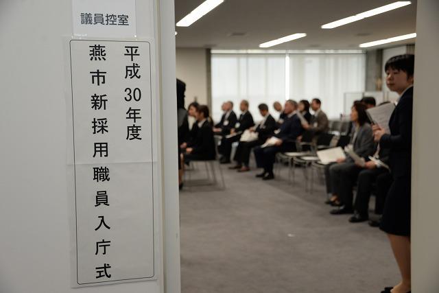 入り口に「平成30年度 燕市新採用職員入庁式」の紙が貼られ、出席者が室内で椅子に座っている写真