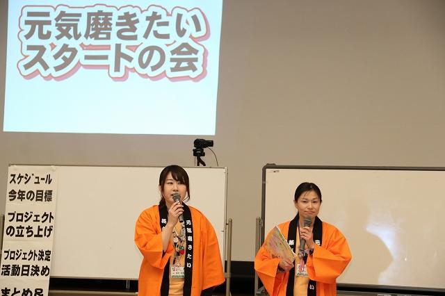 ホワイトボードの前で司会進行しているオレンジ色の法被を着た2人の女性の写真