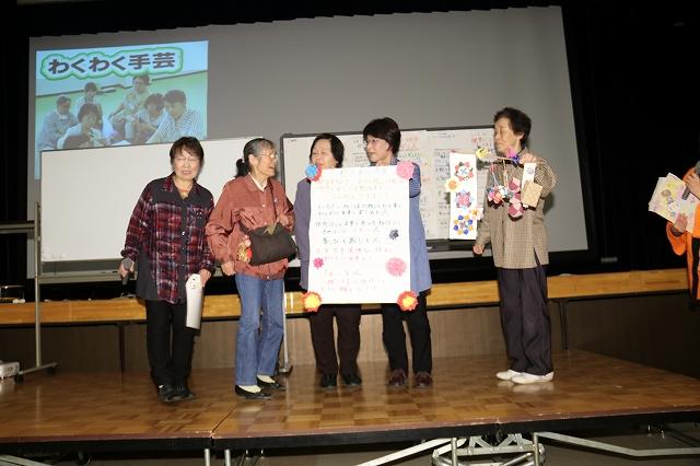 壇上で活動内容と作成した編み物を披露している5人の女性たちの写真