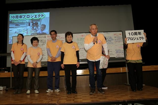 壇上でマイクを片手に活動内容を発表している同じオレンジのTシャツを着た男女6人のグループの様子の写真