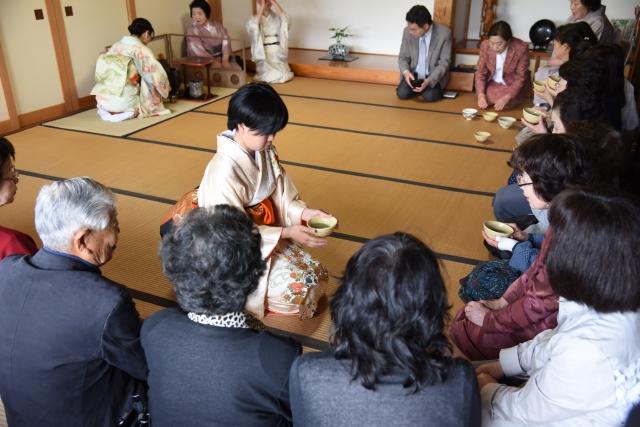 和室で、茶器を持って座っている和服の女性と、周りに座っている参加者たちの写真