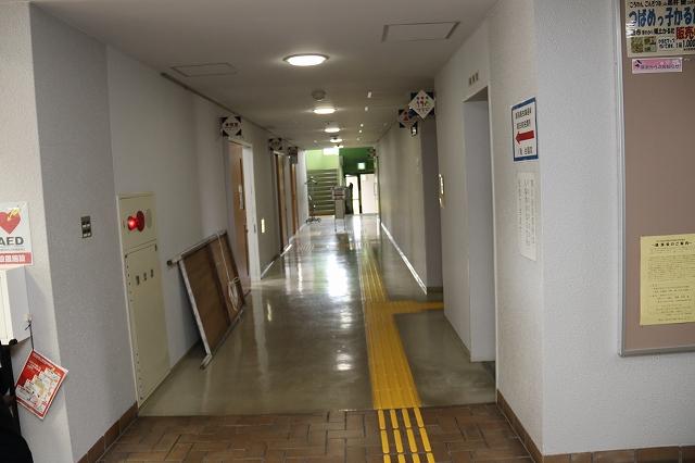 部屋とトイレの出入口に面した公民館の廊下を、奥にある階段に向かって撮影した写真