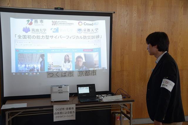 「全国初の総力型サイバーフィジカル防災訓練」のページが表示されたスクリーンと、横で見ているスーツの男性の写真