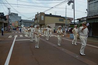 白い制服を着た人たちが、楽器を持って道を行進している写真