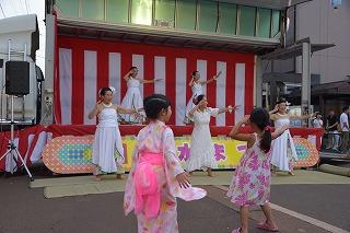 紅白の幕の前で踊っている人たちと、その様子を見ている二人の女の子の写真