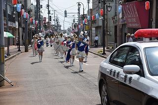 町中で、白いパトカーを先頭に青と白の衣装を着た人たちが並んで行進している写真