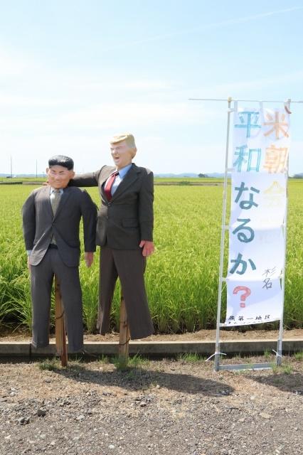 田んぼを背に、灰色のスーツを着た二体のかかしが立っている写真