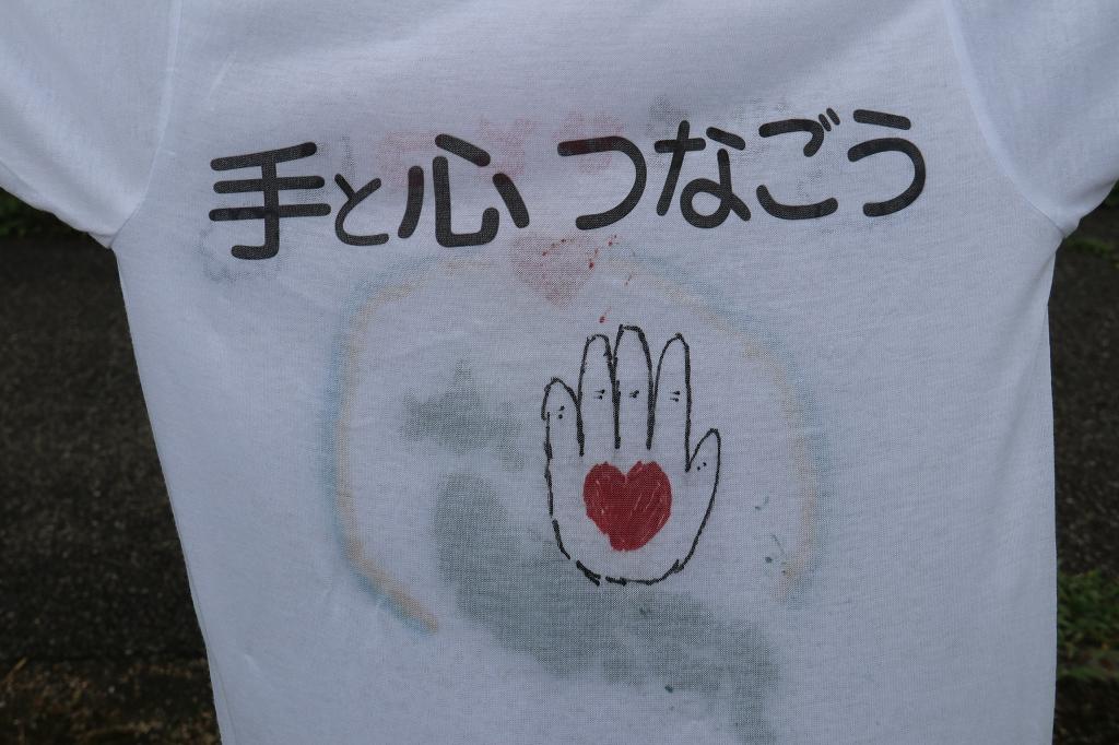「手と心つなごう」という文字の下に、手のひらの中にハートマークが描かれたイラストがあるTシャツの写真