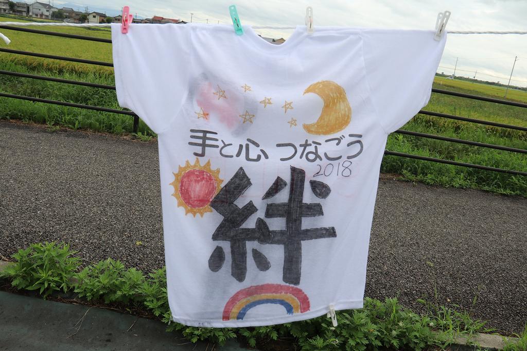 月、太陽、虹などのイラストを背景に、「手と心つなごう 2018 絆」と書かれたTシャツの写真