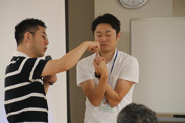 両手をねじったように組んでいる職員と、その両手部分を指差している講師の写真