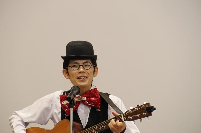 蝶ネクタイの男性が、楽器を演奏しながら歌っている写真