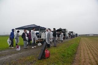 雨合羽を着て田んぼの傍に集まる人たちの写真