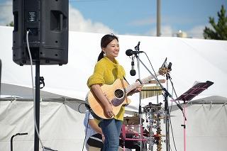 ギターを持ちながら笑顔でマイクに立っている、黄色い服を着た女性の写真