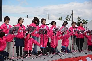 屋外ステージ上でベルなどの楽器を使って演奏をしている、ピンクのジャケットを着た方々の写真