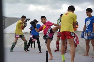 屋外ステージ上でキックボクシング中、左足を蹴り上げているピンクの服の若い男性と、蹴りをガードしている黄色い服の若い男性の写真