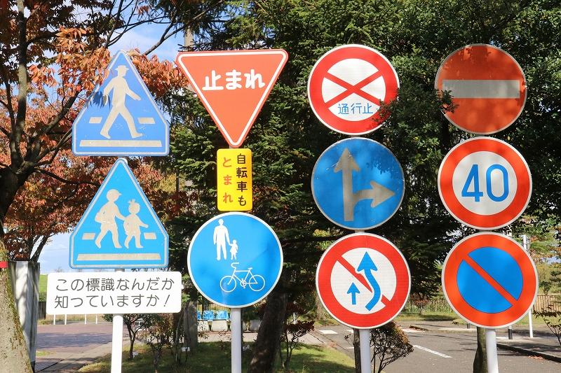 木々を背景にこの標識なんだか知っていますか！と書かれた看板を目印とする自転車と歩行者と自動車に関する11個の交通標識が並ぶ写真