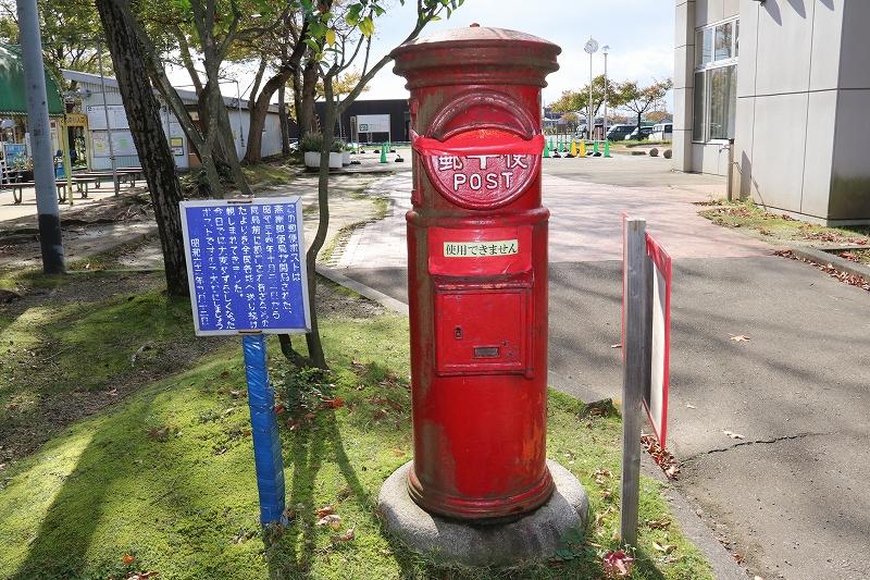 公園内に設置された使用できませんと書かれた丸型の赤い古びた郵便ポストとポストの左脇にあるポストに関する説明書きのある青い立て看板の写真