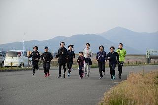 山脈を背景に脇に草が生えたアスファルトの道路の上を駆け抜けるスポーツウエアを着た男女9人の写真