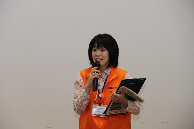 白い壁を背景に黒いクリップファイルと冊子を左手に抱えながら右手に持った黒いマイクで話をするオレンジ色のベストを羽織った女性の写真