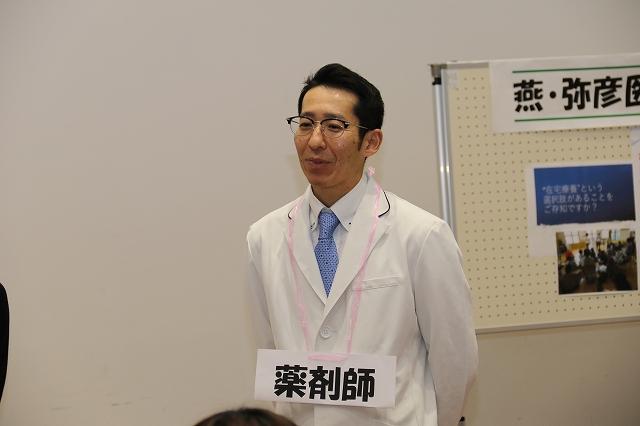 白い壁を背景に後ろで手を組む薬剤師と書かれた下げ札をした白衣を白いシャツの上から着用した眼鏡姿の男性の写真