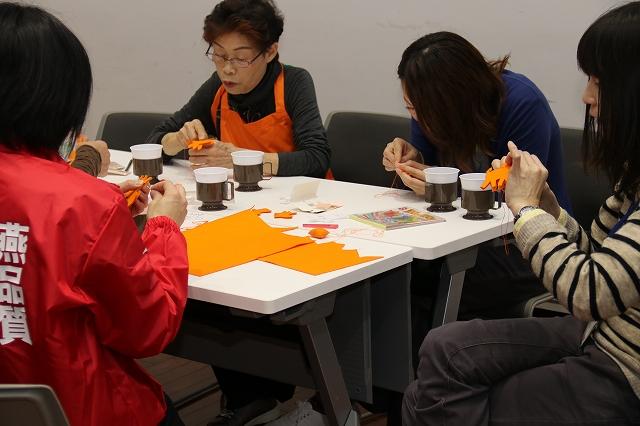5つのコーヒーカップが置かれた白い机の上でロバの形に切り取られた茶色いフェルトを縫い合わせる燕品質を書かれたジャンパーを着た女性とオレンジ色のエプロンを着た女性と青い服を着た女性とボーダー柄のセーターを着た女性の写真