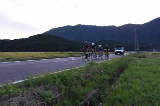 山脈を背景に脇に草の生えた一本の道路を自転車に乗って走る4人の人物と彼らの後ろを走る自動車の写真