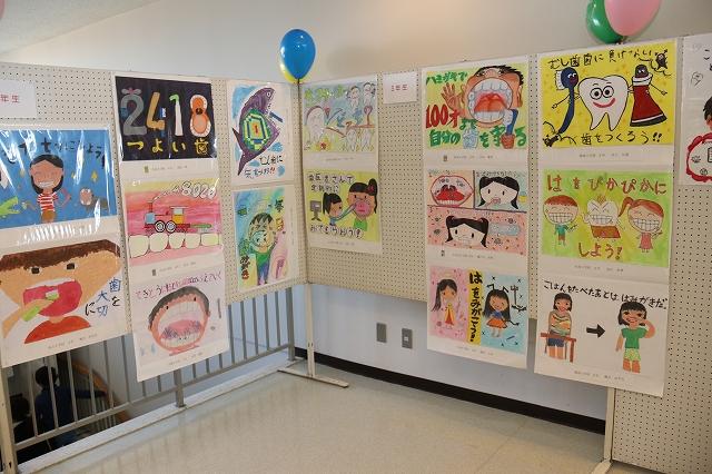 展示された飾りつけの青と黄色・緑色とピンクの2組の4つの風船がついている歯の健康をテーマにした小学校の生徒が描いたイラストが16点展示されたホワイトボードの写真