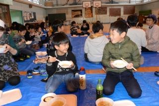 青いマットに座りうれしそうに見つめ合いながら餅を食べている2名の男子生徒と同じく食事をする餅つき大会の参加者の男女生徒や女性たちの写真