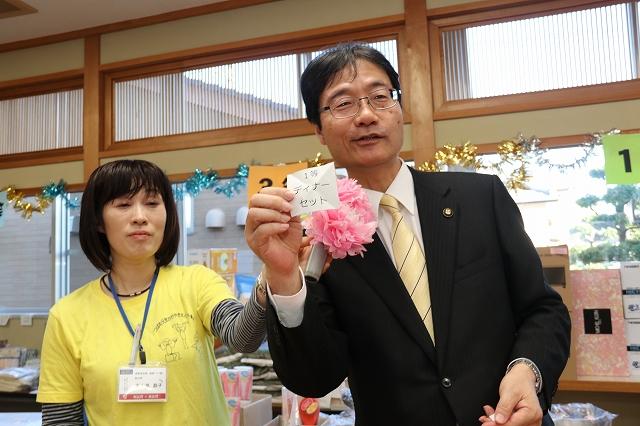 1等ディナーセットと書かれた白いくじを右手に持つスーツ姿の市長と市長の口元に持っているピンク色の花飾りをつけたマイクを近づける黄色いTシャツを着た女性の写真