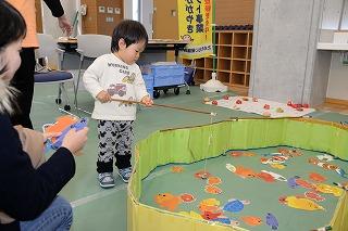床に広げた枠内に紙製の魚をちらばせた魚釣りゲームを置き、実際に体験している幼児の写真