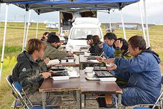 畑の傍でテーブルを設置して食事をしている人たちの写真