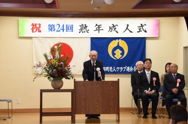 熟年成人式の垂れ幕、日本国旗と老人クラブの旗の前の講壇で謝辞を述べる代表の男性の写真