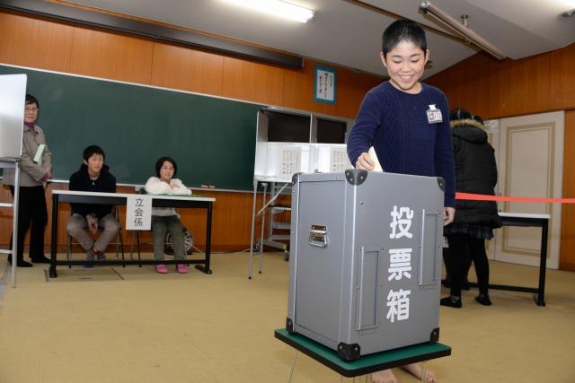 投票箱に選挙用紙を投票する小学生と見守る小学生の立会係の写真