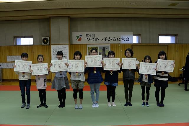 かるた大会会場で、首からメダルを下げ手に賞状を前に掲げて横に並んだ9人の女の子達の写真